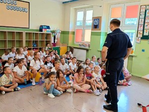 Policjant stoi przed siedzącymi dziećmi i mówi im o zasadach bezpieczeństwa.