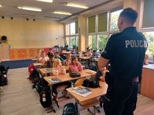 Policjant stoi przed siedzącymi dziecmi i opowiada o bezpieczeństwie.