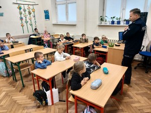 Policjant stoi przed siedzącymi w ławkch dziećmi i opowiada o bezpiecznych zachowaniach na drodze.