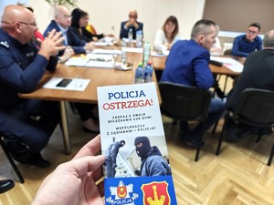 Policjant trzyma ulotkę &quot;Policja Ostrzega!&quot;, w tle widać zebranych gości.