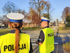 Policjanci stoją przy przejściu dla pieszych, obserwują ruch przy cmentarzu.