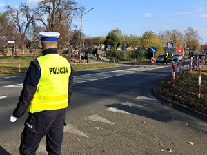 Policjant stoi na skrzyżowaniu i obserwuje ruch w rejonie cmentarza. W tle widać policjanta na przejściu dla pieszych.