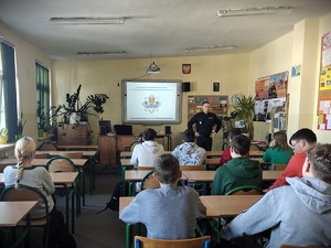 Policjant stoi przed siedzącymi w ławkach szkolnych uczniami.