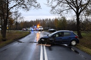 Na pasie jezdni stoi uszkodzony pojazd marki Peugeot. W tle widać pracujące służby.