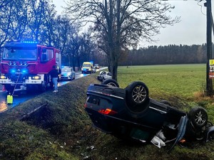 Uszkodzony czarny samochód stoi na dachu w rowie. Na drodze stoją samochody służb ratunkowych.