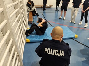 Policjant mierzy czas. Uczennica wykonuje brzuszki leżąc na materacu.