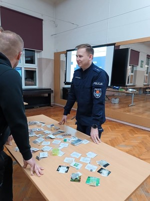 Policjant rozmawia z rodzicami stojącymi przy stoliku. Na stoliku leżą rekwizyty z &quot;walizki narkotykowej&quot;.