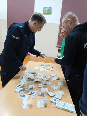 Policjant rozmawia z rodzicami stojącymi przy stoliku, prezentuje zawartość walizki narkotykowej.