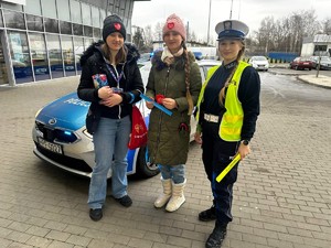 Policjantka pozuje do zdjęcia z dwoma kobietami. W jest radiowóz policyjny.