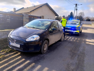 Za czarnym samochodem stoi policyjny radiowóz, który ma włączone światła błyskowe. Pomiędzy samochodami stoi policjant.