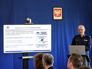 Komendant Powiatowy Policji w Łasku przemawia do zebranych gości. Na ekranie telewizora wyświetla sie prezentacja.