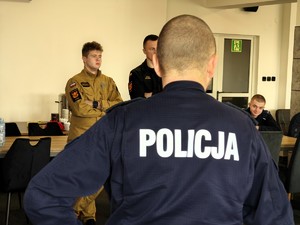 Plecy policjanta przed policjantem stoją strażacy.