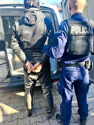 Policjant trzyma pod rękę zatrzymanego mężczyznę. Mężczyzna ma założone kajdanki na ręce trzymane z tyłu.