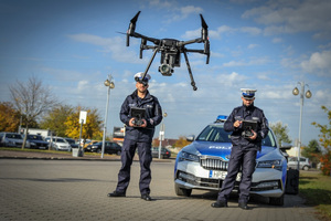 Policjanci stoją przy radiowozie. W powietrze wzbija się dron.