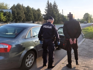 Policjant stoi przy samochodzie razem z mężczyzną w kajdankach.