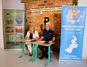 kobieta w jasnej odzieży siedząca po lewej stronie przy stoliku, mężczyzna policjant siedzący przy stoliku po prawej stonie, obok osób ustawione reklamy szkoły oraz policjyjna.