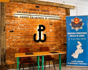 na zdjęciu stolik z dwoma dokumentami. w tle napis Zespół Szkół Mundurowo - Technicznych im. Armii Krajowej w Ostrowie, obok tablica reklamowa z napisem Komenda Powiatowa Policji w Łasku