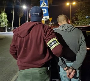 Dwie osoby stojące tyłem. osoba z lewej strony na rękawie posiada opaskę z napisem Policja. Osoba z prawej strony w kajdandkach na rękach z tyłu.