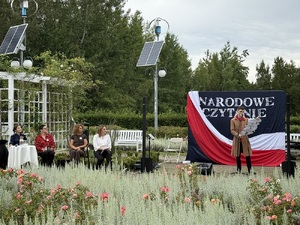 "Narodowe czytanie", widoczny mężczyzna ubrany w starodawny strój czytający tekst. Obok niego siedzące osoby.