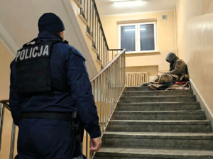 Policjant patrzy na mężczyznę siedzącego na klatce schodowej.
