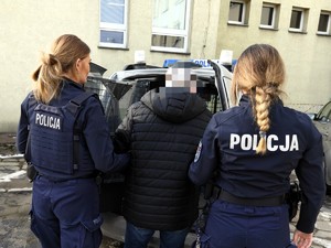 Policjantki trzymają pod rękę poszukiwanego mężczyznę w kajdankach.