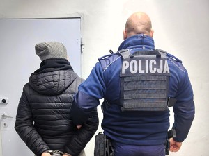 Policjant trzyma pod rękę poszukiwanego mężczyznę w kajdankach.