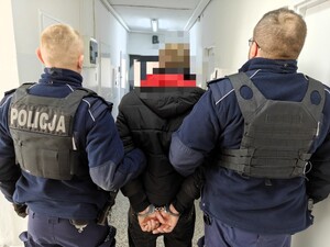 Policjanci trzymają pod rękę poszukiwanego mężczyznę w kajdankach.