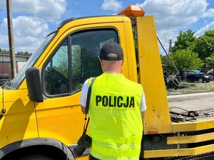 funkcjonariusz Policji w kamizelce odblaskowej, odwrócony tyłem do obiektywu kamery. przed policjantem pojazd typu laweta.