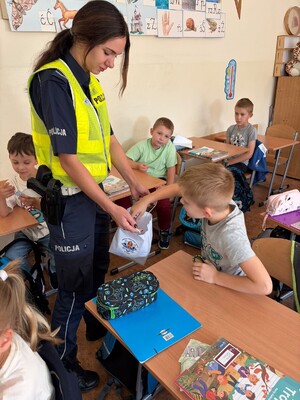 Funkcjonariusza Policji rozdająca dzieciom odblaski. Dzieci siedzą w ławkach.