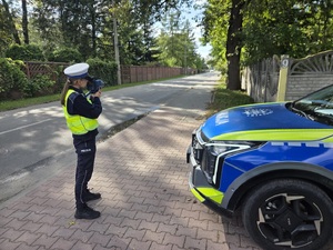 Na zdjęciu policjanta w mundurze trzymająca w rękach miernik prędkości. Obok stojący oznakowany radiowóz.